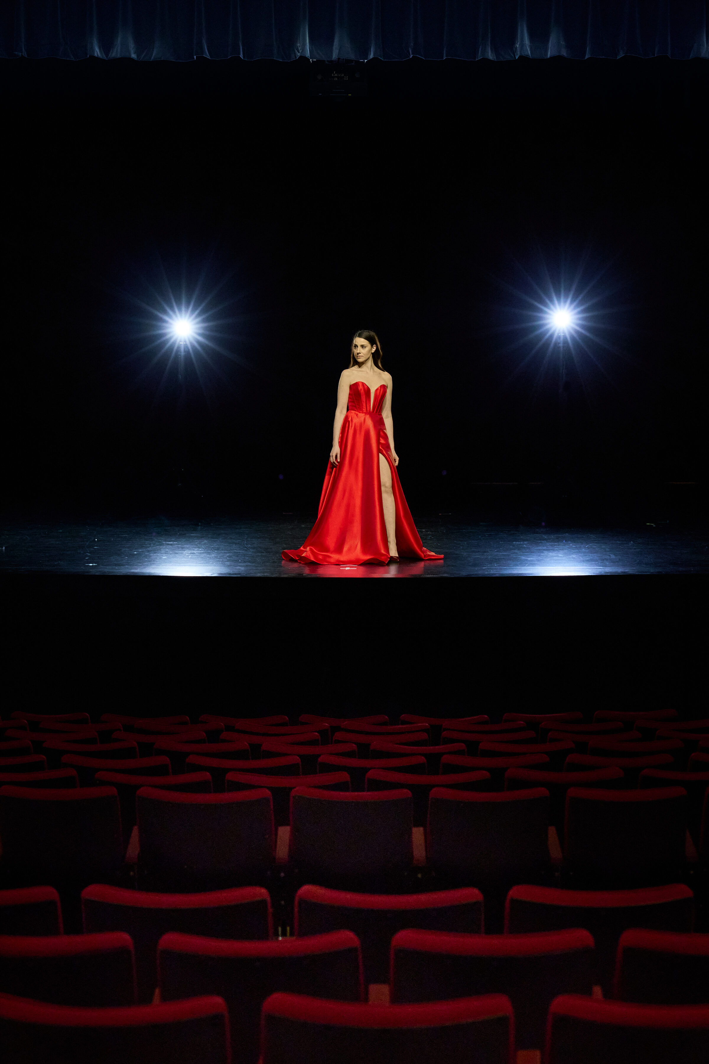 Regina Bianca standing on a theatre stage in a red gown beneath two spotlights